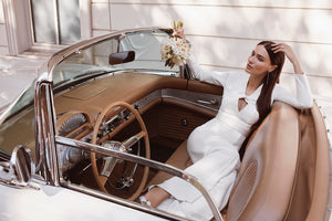 bride sitting in wedding car holding a bouquet wearing a white bridal jumpsuit